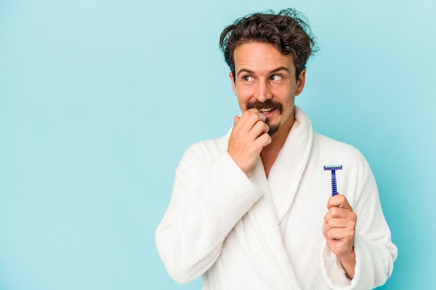 Young caucasian man holding a razor blade isolated on blue background relaxed thinking about something looking at a copy space.