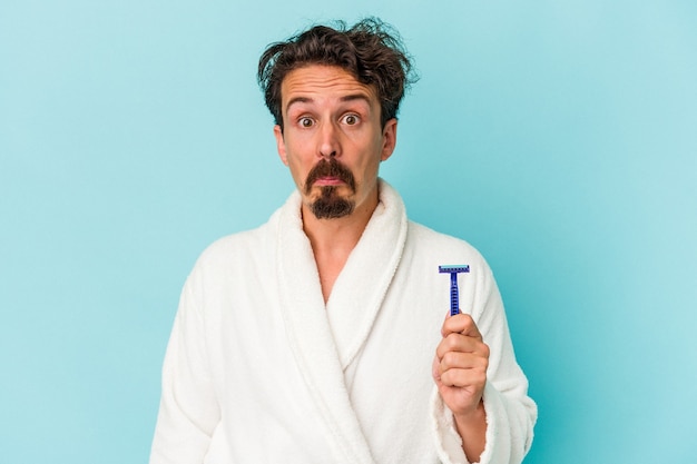 Young caucasian man holding a razor blade isolated on blue background shrugs shoulders and open eyes confused.