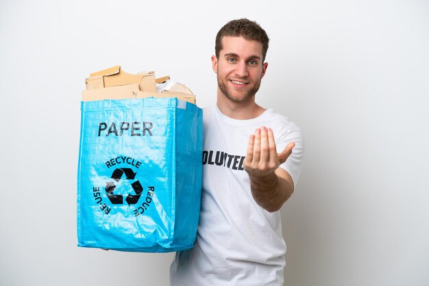 Young caucasian man holding a recycling bag full of paper to recycle isolated on white background inviting to come with hand Happy that you came