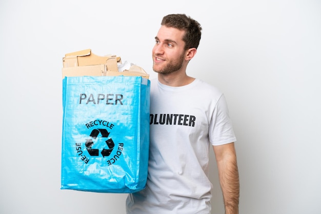 Молодой кавказский мужчина держит мешок для рециркуляции, полный бумаги для переработки, изолирован на белом фоне, смотрит в сторону и улыбается