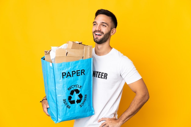 Молодой кавказский мужчина держит мешок для рециркуляции, полный бумаги для переработки, изолирован на белом фоне, позирует с руками на бедрах и улыбается