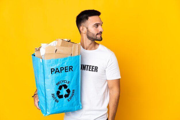 Young caucasian man holding a recycling bag full of paper to recycle isolated on white wall looking to the side