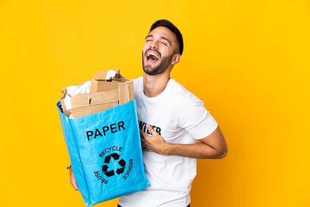 Giovane uomo caucasico che tiene un sacchetto di riciclaggio pieno di carta da riciclare isolato sulla parete bianca che sorride molto