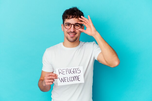 Young caucasian man holding a Refugees welcome placard excited keeping ok gesture on eye.