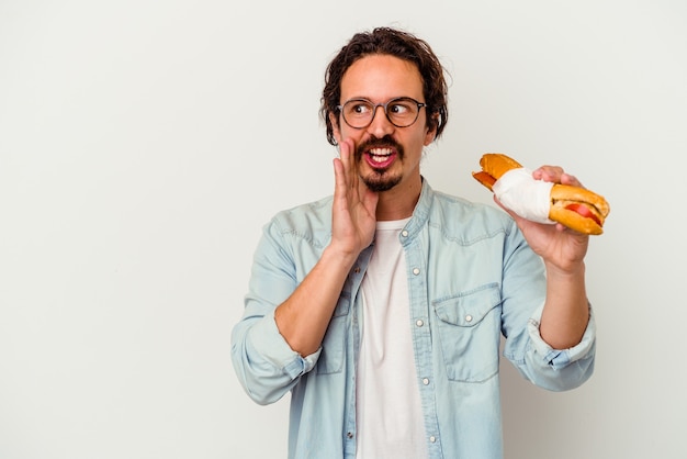 Young caucasian man holding a sandwich isolated on white wall is saying a secret hot braking news and looking aside