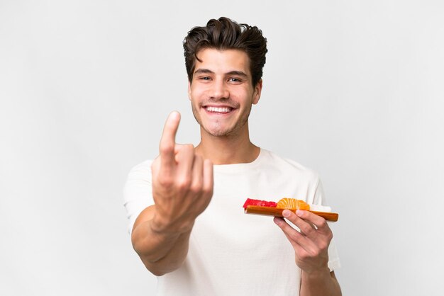 Young caucasian man holding sashimi over isolated white background doing coming gesture