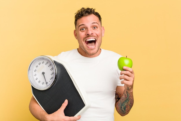 Young caucasian man holding a scale and an apple isolated on yellow background