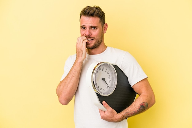 Young caucasian man holding a scale isolated on yellow background biting fingernails, nervous and very anxious.
