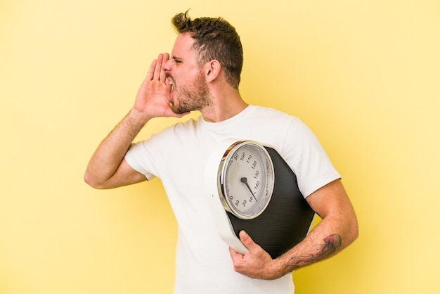 Young caucasian man holding a scale isolated on yellow background shouting and holding palm near opened mouth.