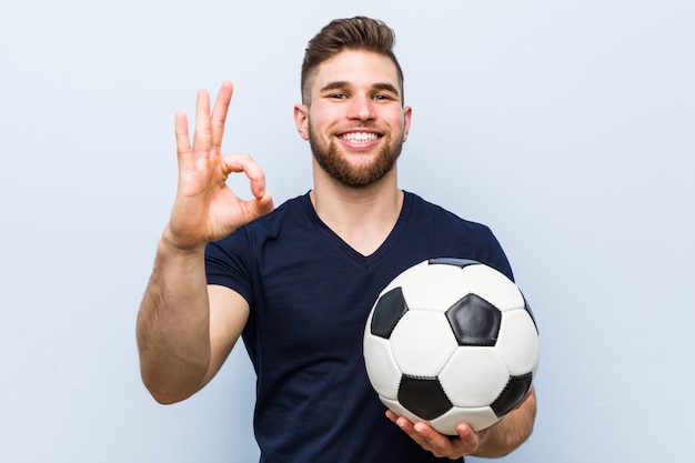 Young caucasian man holding a soccer ball cheerful and confident showing ok gesture.