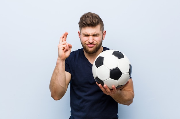 Молодой кавказский человек держа пальцы скрещивания футбольного мяча для иметь удачу