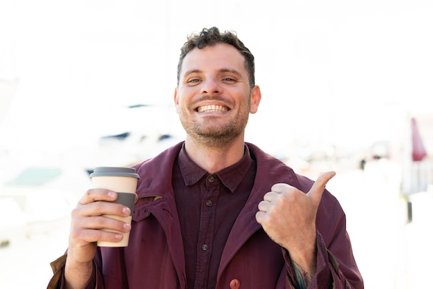 Photo young caucasian man holding a take away coffee at outdoors pointing to the side to present a product