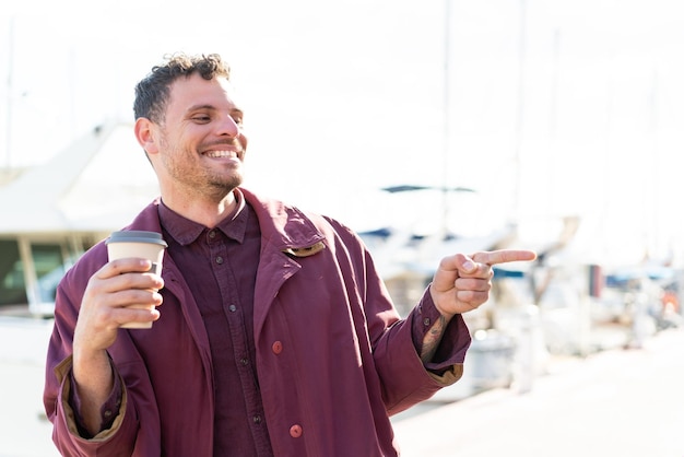 Young caucasian man holding a take away coffee at outdoors pointing to the side to present a product