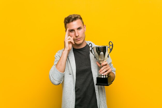 Young caucasian man holding a trophy pointing his temple with finger, thinking, focused on task.