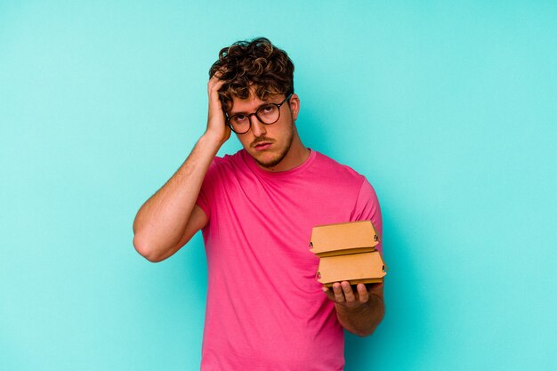 Young caucasian man holding two burgers isolated on blue background being shocked, she has remembered important meeting.