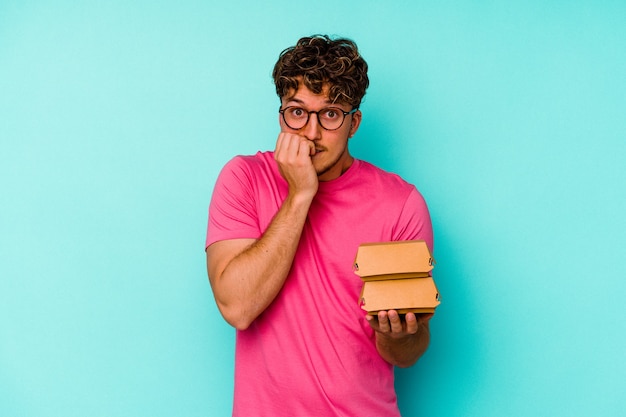 Young caucasian man holding two burgers isolated on blue background biting fingernails, nervous and very anxious.