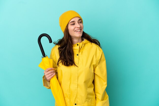 Young caucasian man holding an umbrella isolated on blue background looking to the side and smiling