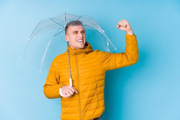 Young caucasian man holding an umbrella raising fist after a victory, winner concept.