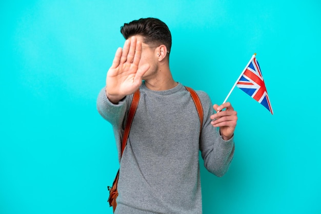 Young caucasian man holding an United Kingdom flag isolated on blue background making stop gesture and disappointed