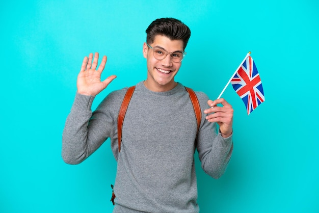 Young caucasian man holding an United Kingdom flag isolated on blue background saluting with hand with happy expression