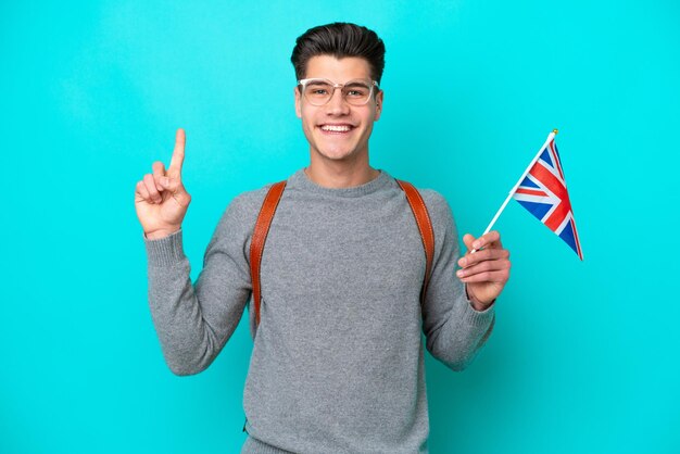 Young caucasian man holding an United Kingdom flag isolated on blue background showing and lifting a finger in sign of the best