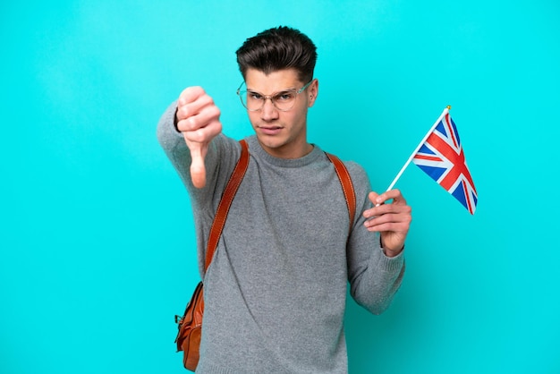 Young caucasian man holding an United Kingdom flag isolated on blue background showing thumb down with negative expression