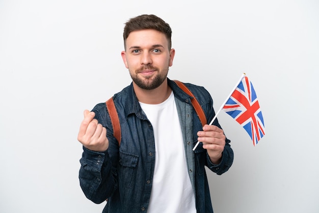 Young caucasian man holding an United Kingdom flag isolated on white background making money gesture