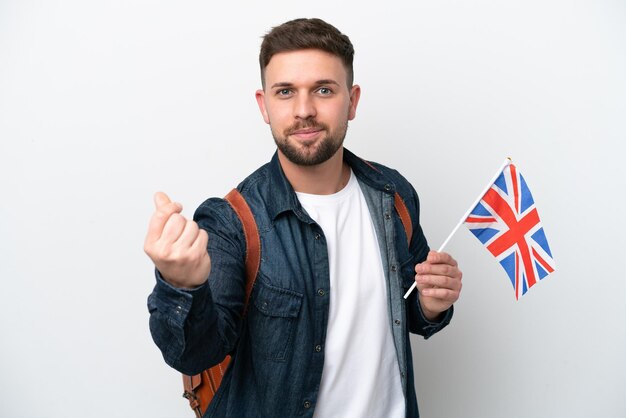 Young caucasian man holding an United Kingdom flag isolated on white background making money gesture