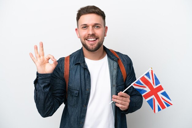 손가락으로 확인 표시를 보여주는 흰색 배경에 고립 된 영국 국기를 들고 젊은 백인 남자