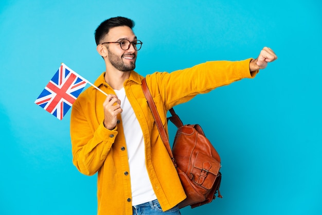 Young caucasian man holding an United Kingdom flag isolated on yellow background giving a thumbs up gesture