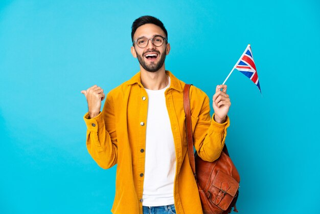 Young caucasian man holding an United Kingdom flag isolated on yellow wall pointing to the side to present a product