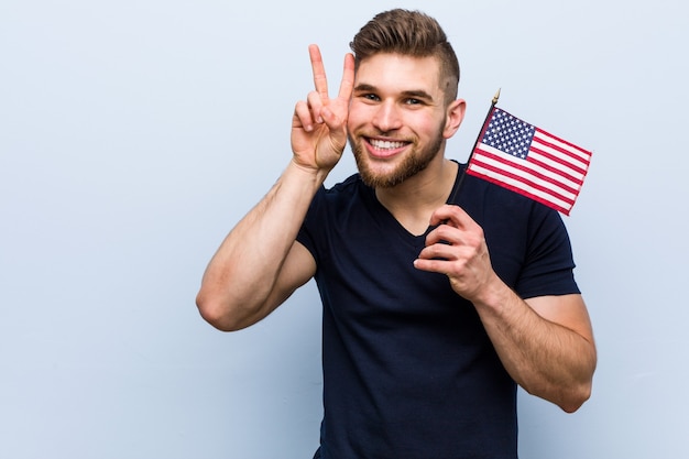 Молодой кавказский мужчина держит флаг Соединенных Штатов, показывая знак победы и широко улыбается.
