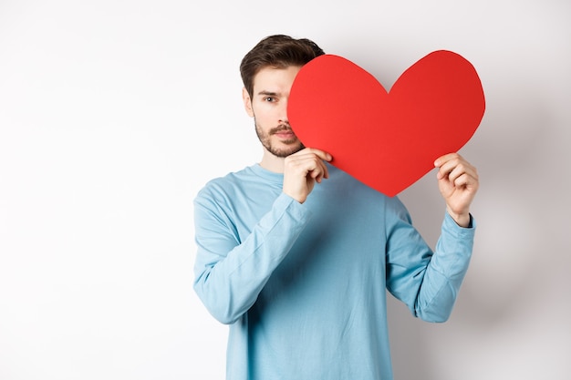 Young caucasian man holding Valentines day romantic heart, con half of face with cutout and smiling, searching for love date on white