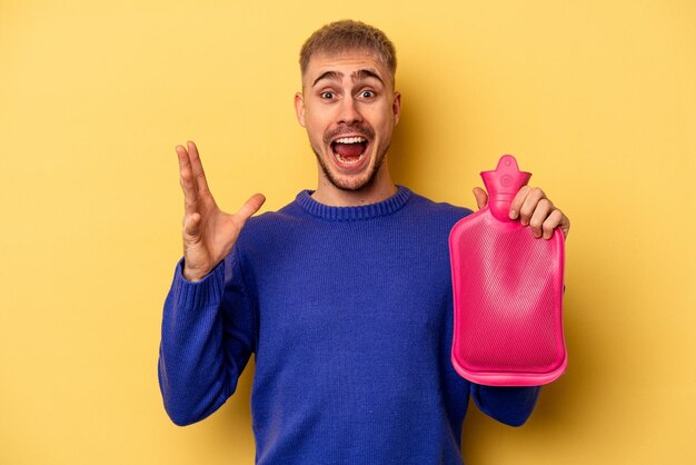 Giovane uomo caucasico che tiene una sacca d'acqua isolata su sfondo giallo che riceve una piacevole sorpresa, eccitato e alzando le mani.