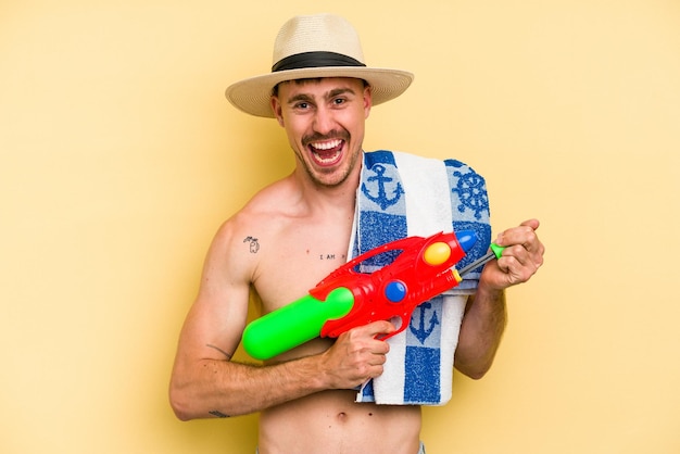 Young caucasian man holding water gun isolated on yellow background