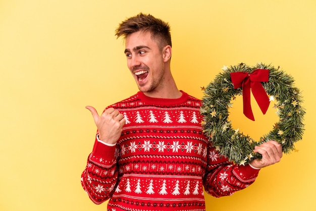 Young caucasian man holding a wreath isolated on yellow background points with thumb finger away, laughing and carefree.