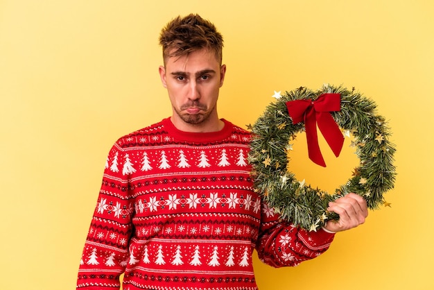 Young caucasian man holding a wreath isolated on yellow background shrugs shoulders and open eyes confused.