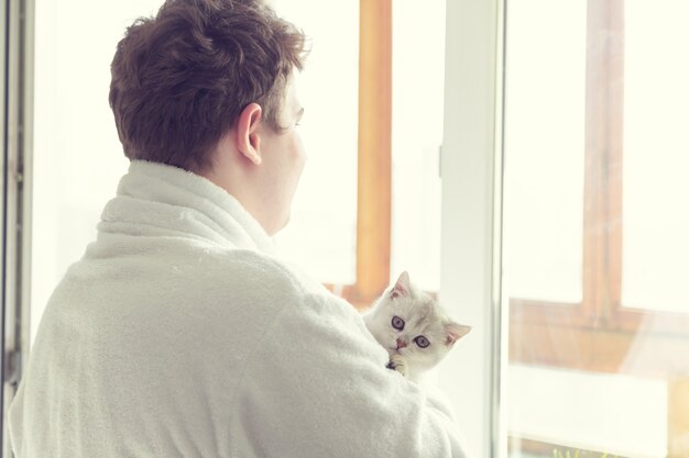 Young caucasian man is drinking coffee near the window. He is holding a cat. Early morning, sunshine.