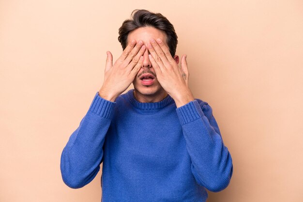 Young caucasian man isolated on beige background afraid covering eyes with hands.