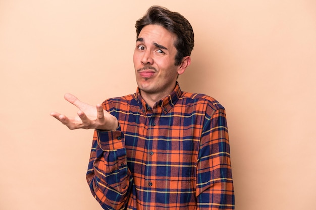 Young caucasian man isolated on beige background doubting and shrugging shoulders in questioning gesture.