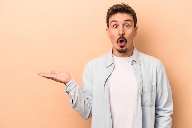 Young caucasian man isolated on beige background impressed holding copy space on palm