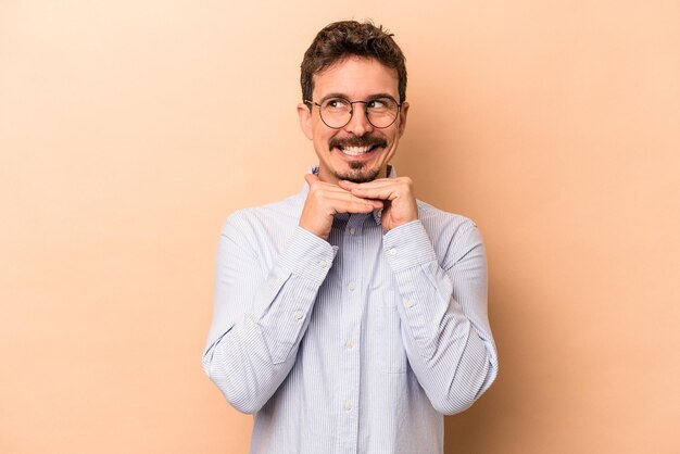 Young caucasian man isolated on beige background keeps hands under chin, is looking happily aside.
