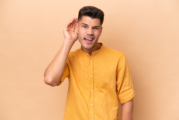 Young caucasian man isolated on beige background listening to something by putting hand on the ear