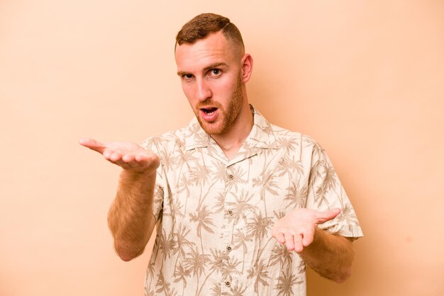 Young caucasian man isolated on beige background makes scale with arms feels happy and confident