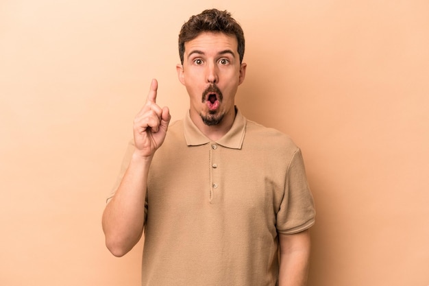 Young caucasian man isolated on beige background pointing upside with opened mouth.