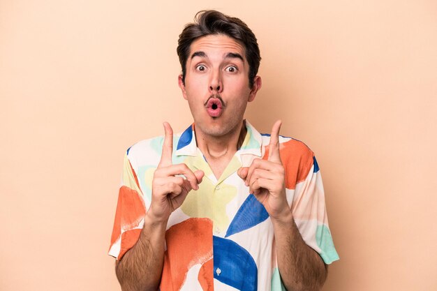 Young caucasian man isolated on beige background pointing upside with opened mouth