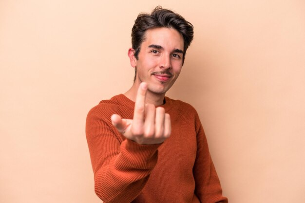 Young caucasian man isolated on beige background pointing with finger at you as if inviting come closer.