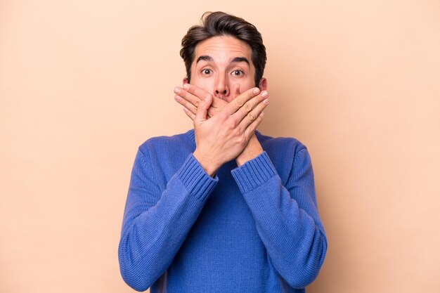Young caucasian man isolated on beige background shocked covering mouth with hands.