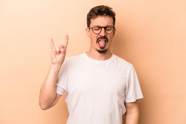 Young caucasian man isolated on beige background showing rock gesture with fingers