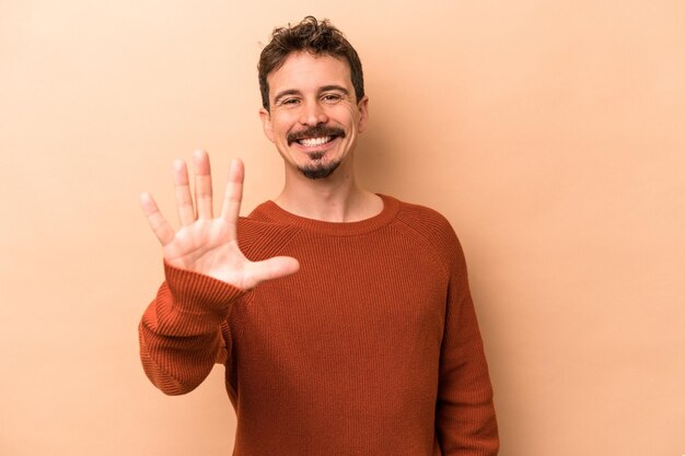 Young caucasian man isolated on beige background smiling cheerful showing number five with fingers.